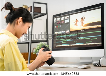 Asian woman holding a camera and using application video editor works on the computer with footage on wooden table.