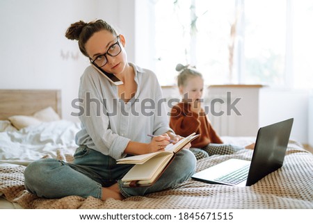 Young mother talking on mobile phone and working on a laptop. Business Woman working from home with little girl. Work on maternity leave. Covid-19.