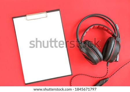 Black headphones and clipboard notepad with white blank paper on red background. Concept listening to audio material and abstract, distance education. Top view copy space.
