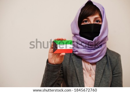 Portrait of young muslim woman wearing formal wear, protect face mask and hijab head scarf, hold Kuwait flag card against isolated background. Coronavirus country concept.