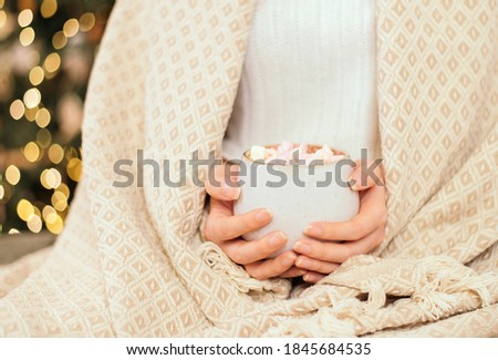 A girl wrapped in a plaid in a knitted white woolen sweater holds a mug with hot chocolate or coffee with marshmallows. Christmas lights on, cozy holiday atmosphere, copy space. Close up. noise effect