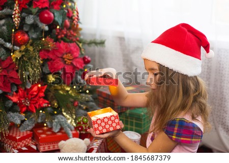 Little girl in Santa hat opens a red box with a gift and a Golden magic light near the Christmas tree. Holiday decor, poinsettias on fir trees, New year. Joy, surprise, children's emotions. Copyspace