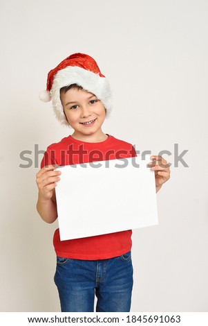 Portrait of a beautiful little boy in a red hat and red t-shirt on a white background. A child in a Christmas hat of Santa Claus is holding a clean white sheet of paper.