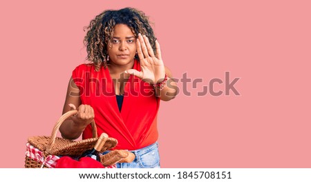 Young african american plus size woman holding picnic wicker basket with bread with open hand doing stop sign with serious and confident expression, defense gesture 