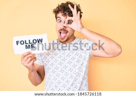 Young caucasian man with curly hair holding follow me message paper smiling happy doing ok sign with hand on eye looking through fingers 