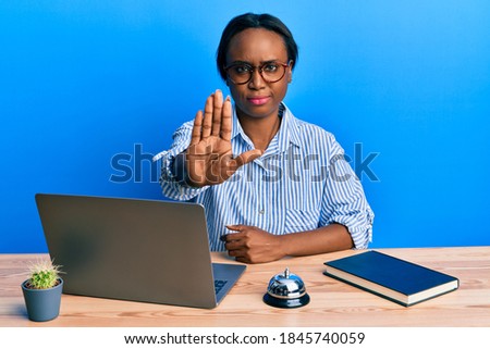 Young african woman working at hotel reception using laptop doing stop sing with palm of the hand. warning expression with negative and serious gesture on the face. 