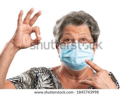 Elderly female wearing glasses or spectacles pointing index finger at disposable medical mask to prevent covid19 influenza virus infection making okay gesture using hand isolated on white