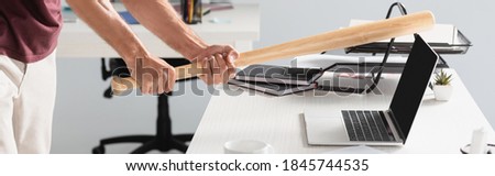 Cropped view of businessman beating laptop with baseball bat with blurred background in office, banner