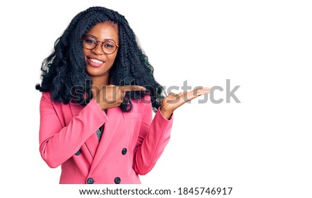 Beautiful african american woman wearing business jacket and glasses amazed and smiling to the camera while presenting with hand and pointing with finger. 