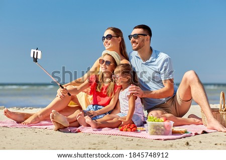 family, leisure and people concept - father, mother and two little daughters taking picture with smartphone and selfie stick on summer beach