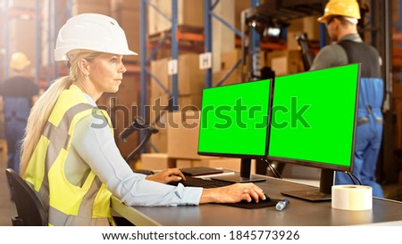 Professional Female Worker Wearing Hard Hat Uses Computer with Green Chroma Screen Mock-up in the Retail Warehouse full of Shelves with Goods. Distribution Logistical Center