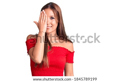 Young beautiful hispanic woman wearing casual clothes covering one eye with hand, confident smile on face and surprise emotion. 