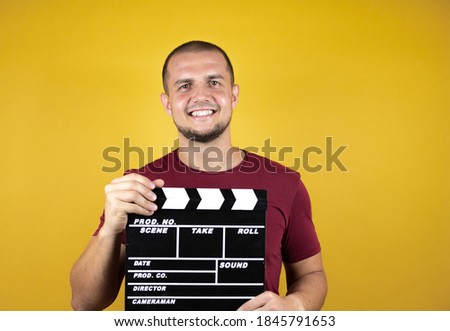 Russian man wearing basic red t-shirt over yellow insolated background standing over isolated yellow background holding clapperboard very happy