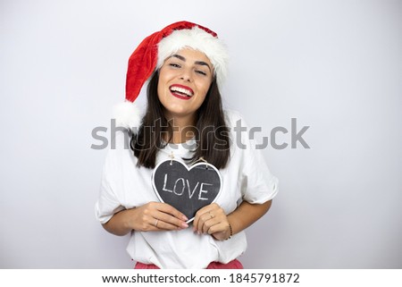 Young beautiful woman wearing a christmas hat over white background holding blackboard with love word message smiling looking at the camera