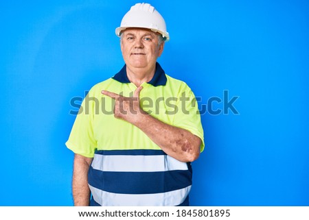 Senior grey-haired man wearing worker reflective t shirt and hardhat smiling cheerful pointing with hand and finger up to the side 