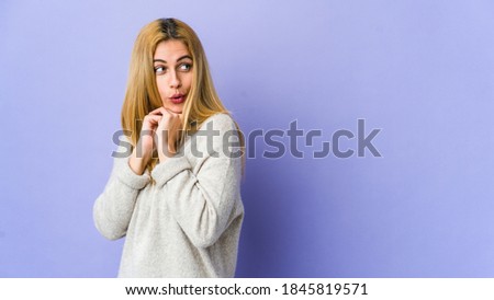 Young blonde woman isolated on purple background praying for luck, amazed and opening mouth looking to front.