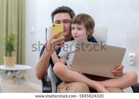 Happy young father sit on chair using laptop relax with kid son holding smartphone have fun together, smiling dad and little boy child enjoy weekend at home rest sofa busy with gadgets and play games.
