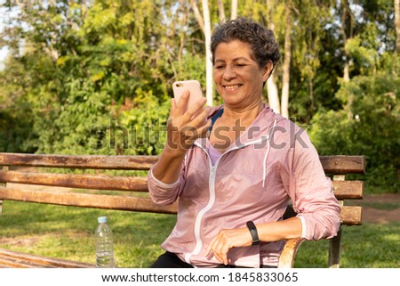 Old aged brazilian woman looking mobile screen to use app. Outdoors in sunny day. Network, messaging, internet, lifestyle concept.