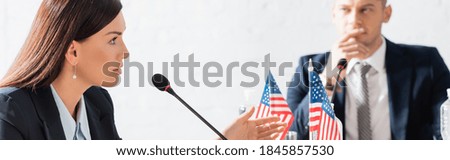Female politician gesturing, while speaking in microphone, sitting near american flags with blurred man on blurred background, banner