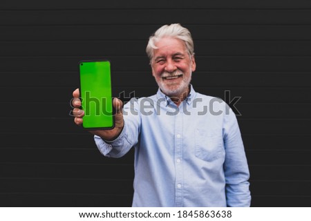 old and mature man holding his smartphone showing it at the camera with the green screen - smiling happy adult portrait using technology