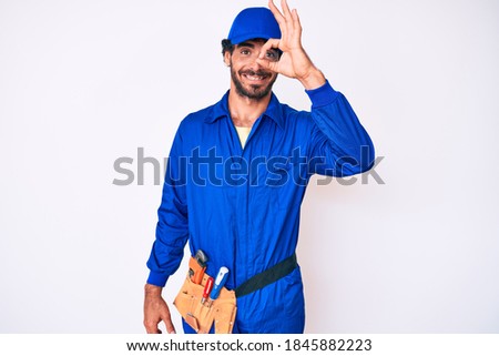 Handsome young man with curly hair and bear weaing handyman uniform doing ok gesture with hand smiling, eye looking through fingers with happy face. 