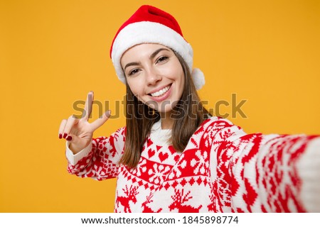 Close up of smiling young Santa woman in sweater Christmas hat doing selfie shot on mobile phone showing victory sign isolated on yellow background. Happy New Year celebration merry holiday concept