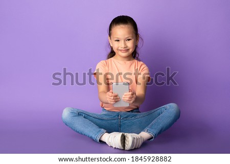 Childhood And Technology. Smiling asian girl holding and using smartphone, browsing internet, watching video or cartoons, sitting on the floor over pastel purple background , copy space