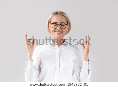 Stress management concept. Calm mature business lady in office wear meditating on light grey studio background. Tranquil female secretary feeling relaxed and balanced, practicing workplace yoga