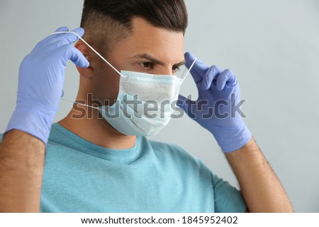 Man in medical gloves putting on protective face mask against grey background