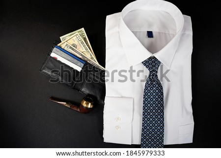 folded white dress shirt with necktie and money in black leather wallet and pipe on black background