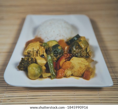A portion of yellow rice curry with chicken and fresh organic vegetables on a white plate, product picture with curry sauce, chili dip, ginger and basil