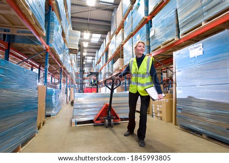 A wide shot of a female warehouse manager holding a clipboard and pulling a pallet truck. Royalty-Free Stock Photo #1845993805