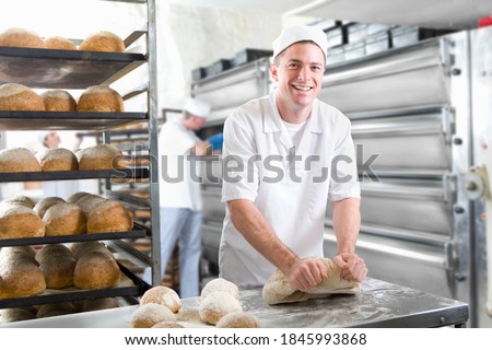 A medium shot of a young baker smiling and looking the camera while kneading bread dough in a bakery. Royalty-Free Stock Photo #1845993868