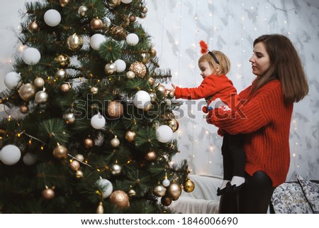 Mom and daughter in red warm sweaters decorate the Christmas tree. Happy motherhood. Warm family relationships. Christmas and New Year's interior. Love. Family concept.