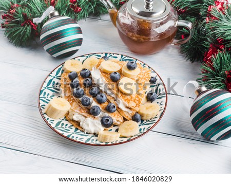 Christmas tree shaped sweet crepes filled with cream greek yogurt, banana and blueberries on white wooden table with winter holiday decor.