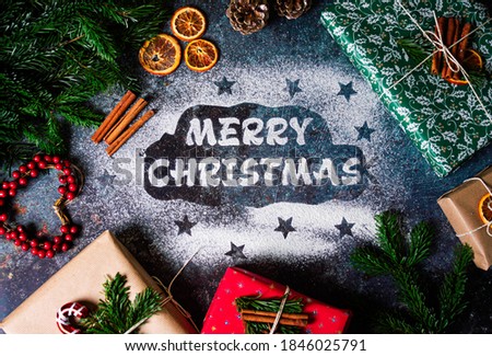 Handmade inscription Merry Christmas from flour on dark background with gift boxes in colorful wrapping paper, dry oranges, pine branch, cinnamon. Holiday postcard. View from above. Art photo.  
