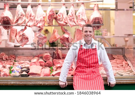 A young smiling butcher in a red apron standing and leaning his back against the butcher shop window. Royalty-Free Stock Photo #1846038964