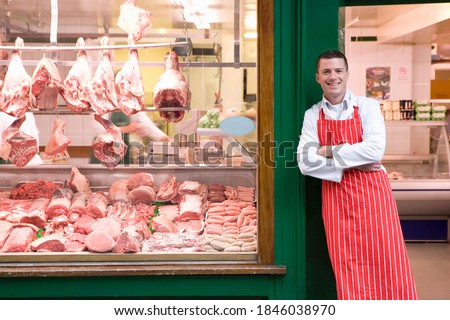 A young smiling butcher in red apron leaning against butcher shop doorway next to the display window. Royalty-Free Stock Photo #1846038970