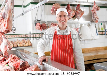 A young smiling butcher wearing a red apron standing next to a meat display case. Royalty-Free Stock Photo #1846038997
