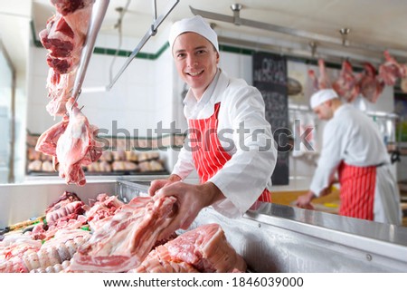 A young butcher smiling and holding meat near the camera in a meat shop. Royalty-Free Stock Photo #1846039000