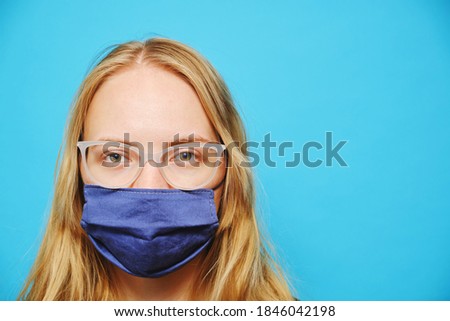 Young blonde Caucasian girl with glasses in a mask on a blue background. Studio photo and copy space.