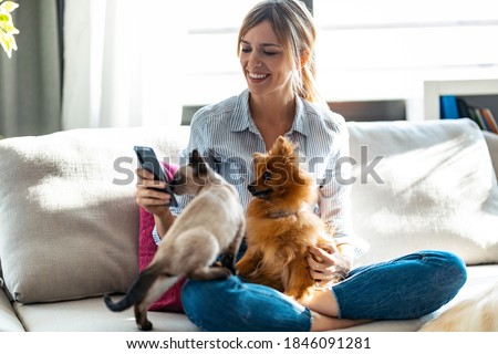 Shot of beautiful young woman playing with her cute dog and cat while using mobile phone sitting on couch in living room at home. Royalty-Free Stock Photo #1846091281