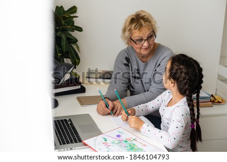 Mature grandmother helping child with homework at home. Satisfied old grandma helping her granddaughter studying in living room. Little girl writing on notebook with senior teacher sitting next to her