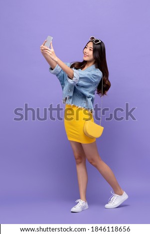 Full length portrait of smiling young pretty Asian woman taking selfie with smartphone in isolated studio purple background 