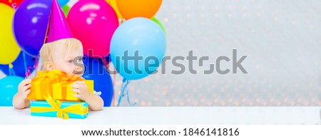 A little girl is sitting in a festive hat on a background of colorful balloons next to a gift and looks at the camera with a smile. First birthday concept. Stretched panoramic image for banner