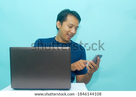 Young Asian man feeling happy and smile what he see in the smartphone and laptop on the table. Indonesia Man wear blue navy t-shirt Isolated blue background.