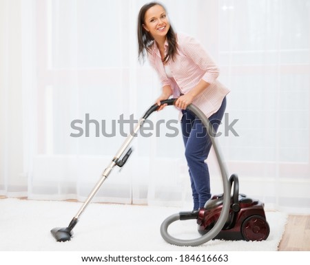 Young cheerful brunette woman vacuum cleaning carpet in home interior 