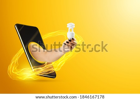 A hand feeds an energy-saving lamp through a smartphone screen on a yellow background. Green technology concept, clean energy, evolution, cool idea