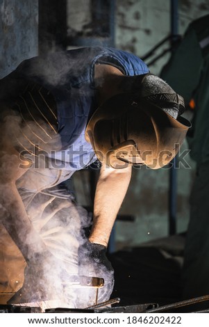 Close up of a strong man is a welder in uniform, welding mask and welders leathers, a metal product is welded with  welding machine at the construction site