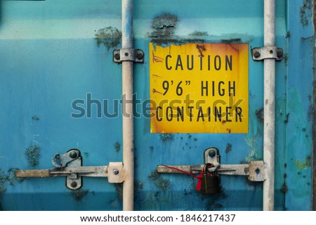 The back door of blue heavy-duty container with locks and sealed padlock. Container is very rusty and aged. Yellow warning sign about high container height is marked on the door